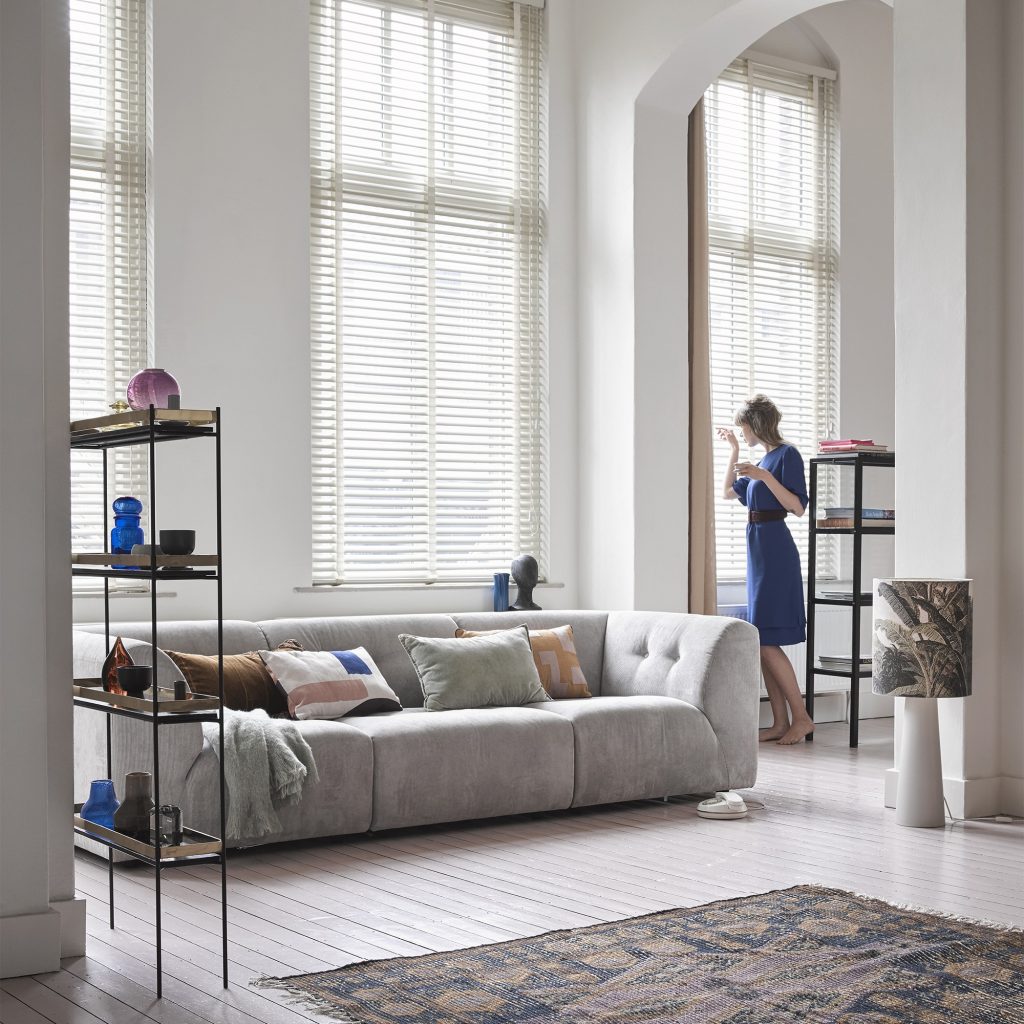 Mujer en salón moderno luminoso con grandes ventanales, sofá gris, estantería metálica y decoración